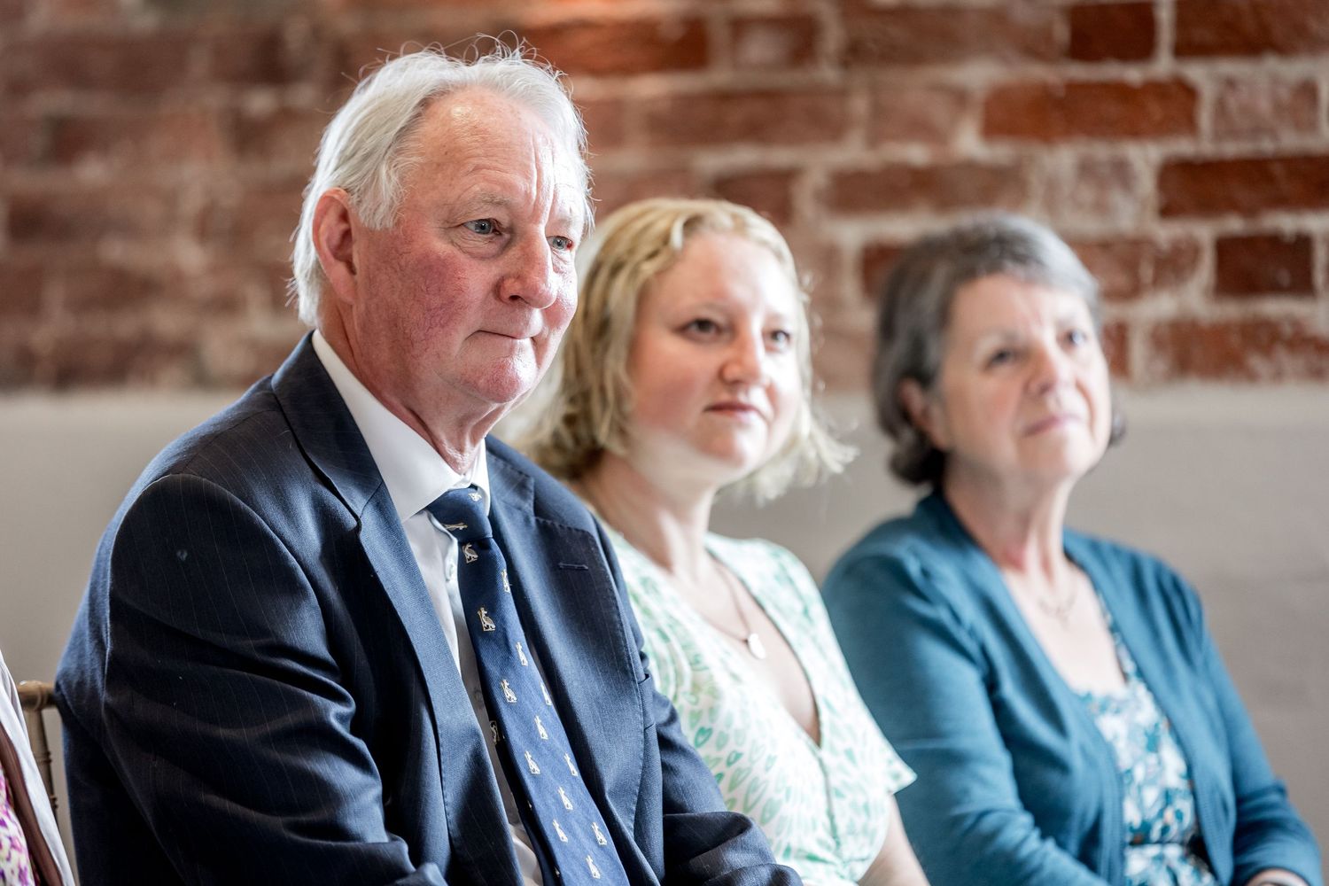 Wedding guests seated in formal attire against a rustic brick wall background.