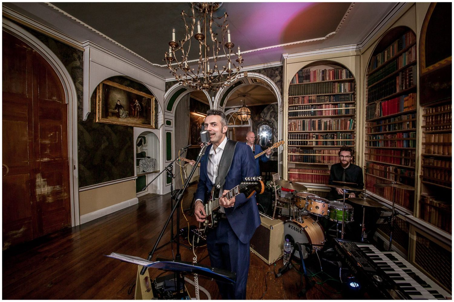 Wedding band performs in an elegant library room with bookshelves and chandelier.