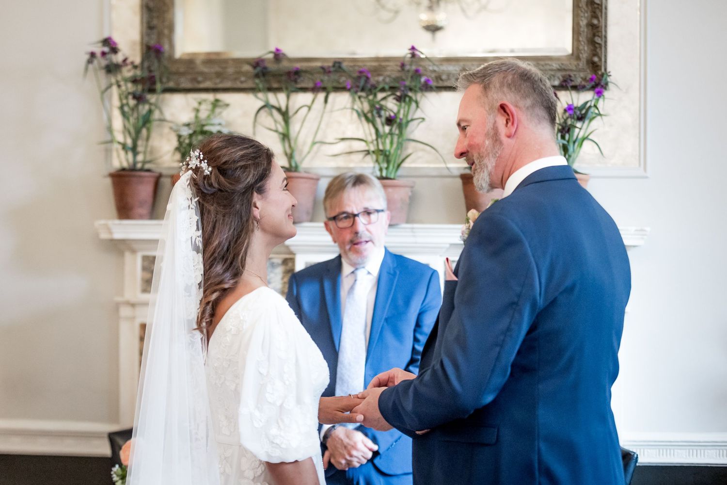 A wedding ceremony in progress in an elegant room with a fireplace and potted plants.