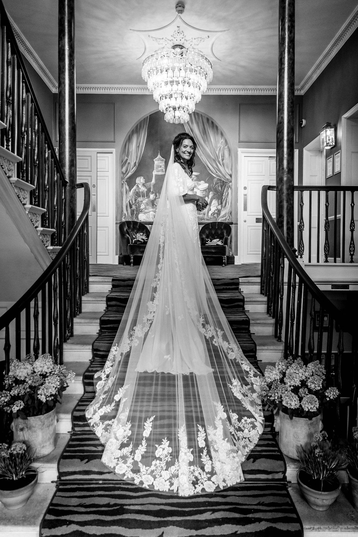 Elegant bridal gown with flowing lace veil photographed on grand staircase with crystal chandelier in black and white.