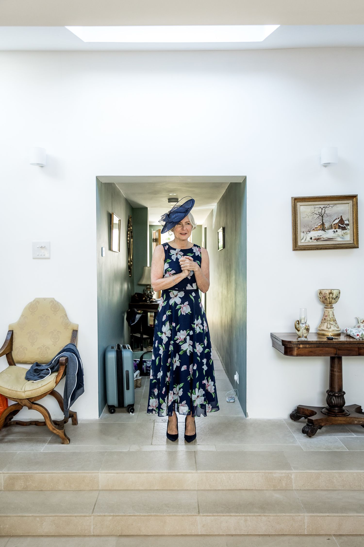 A woman wearing a navy floral dress and blue fascinator stands in an elegant hallway with antique furniture.