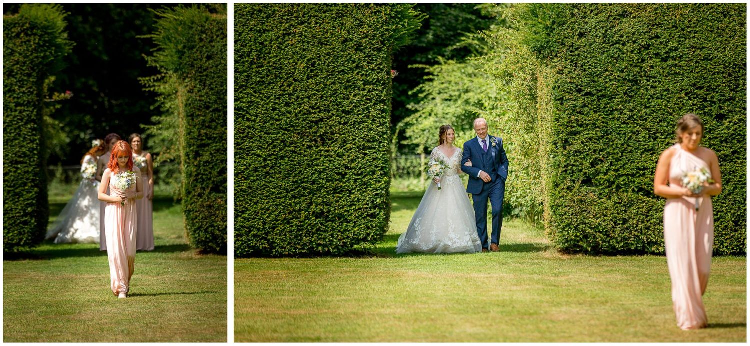 Wedding party members walk and pose on lush green lawn near tall garden hedges during outdoor celebration.