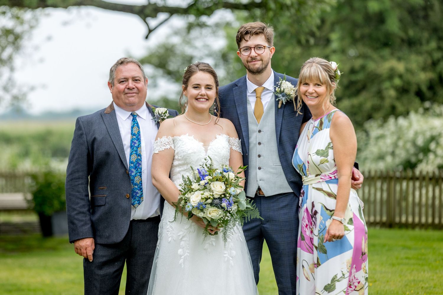 Wedding party members pose together outdoors in formal attire against a green garden backdrop.