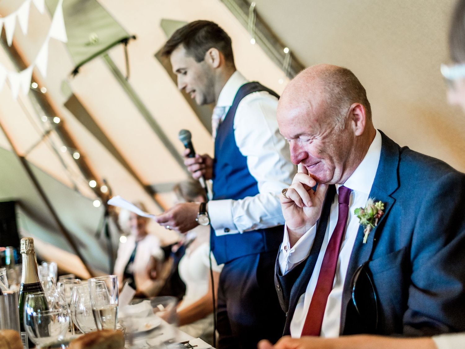 Wedding guests laugh and react emotionally during speeches at an indoor reception venue.