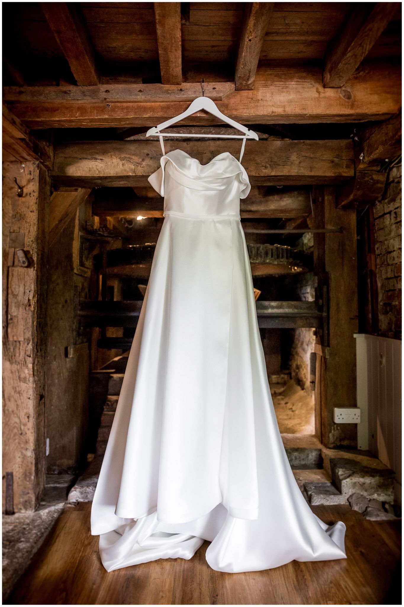 White wedding dress hanging in rustic wooden barn interior with exposed beams.