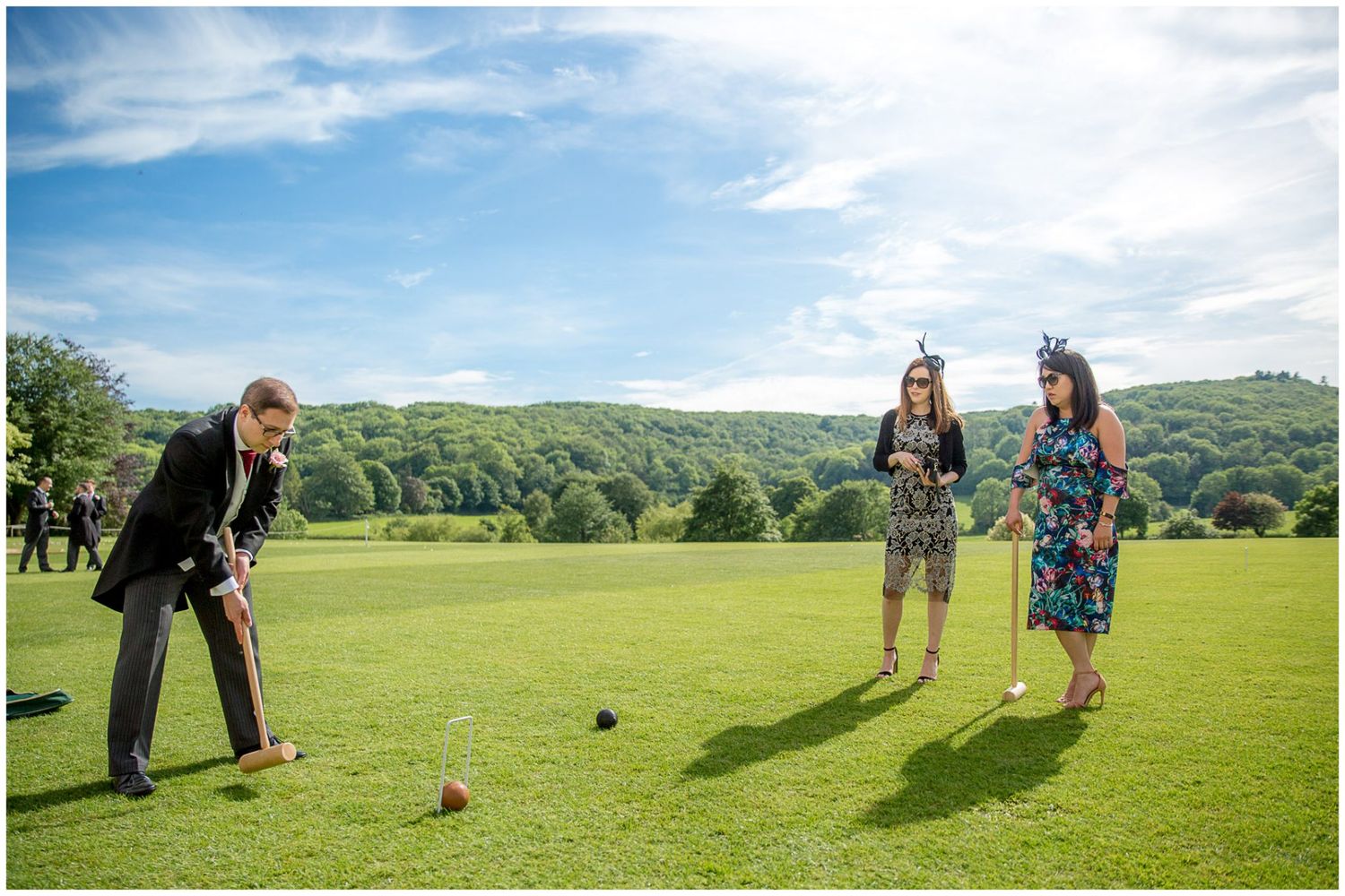People playing croquet on a lush green lawn with rolling hills and blue sky in the background.