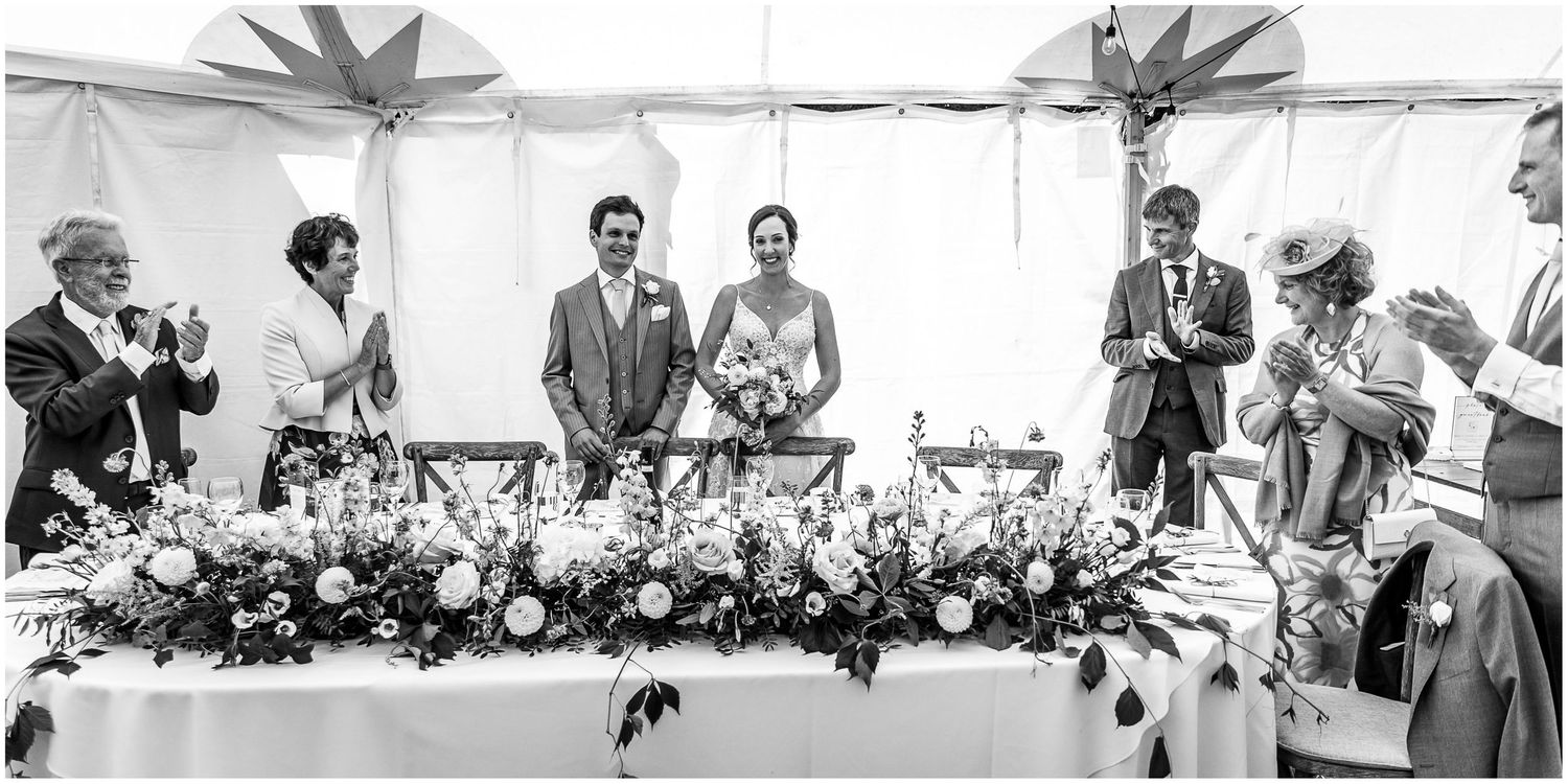 Wedding party gathered at a decorated head table with floral arrangements.
