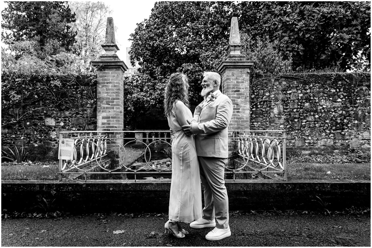 in Winchester, a black and white wedding photo showing a bride and groom posing in front of a stone wall in a garden setting.