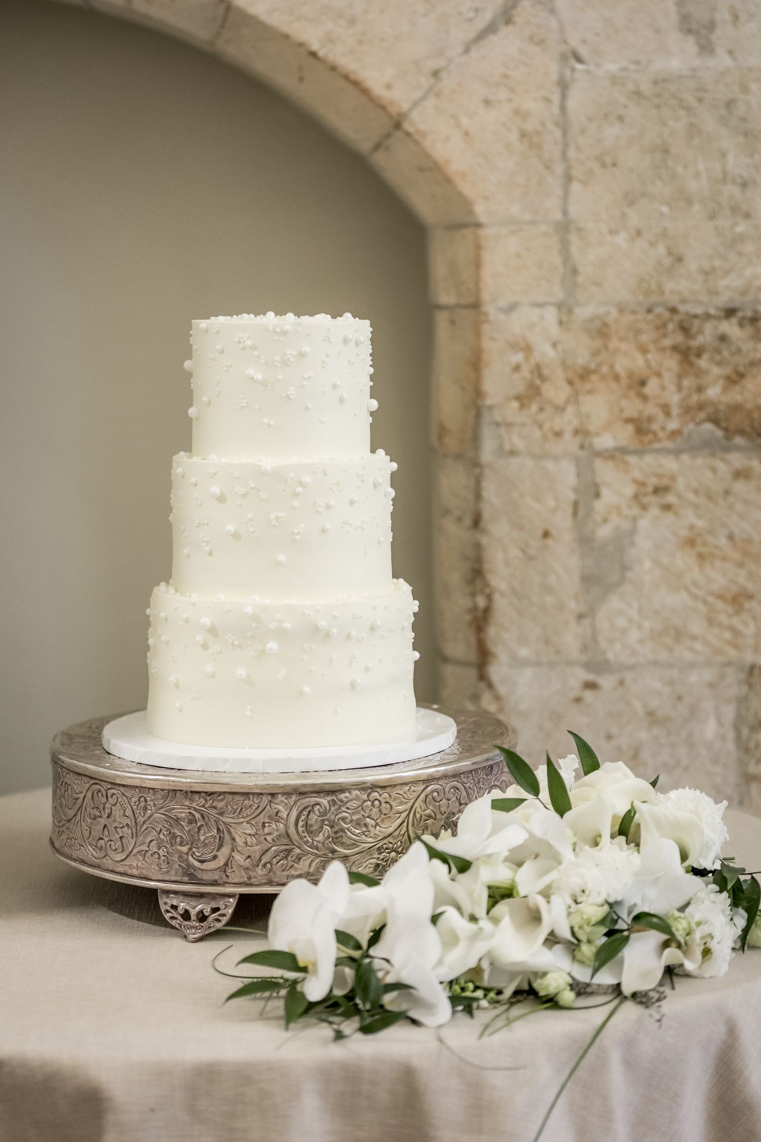 Elegant white tiered wedding cake displayed with white floral decorations against stone archway.