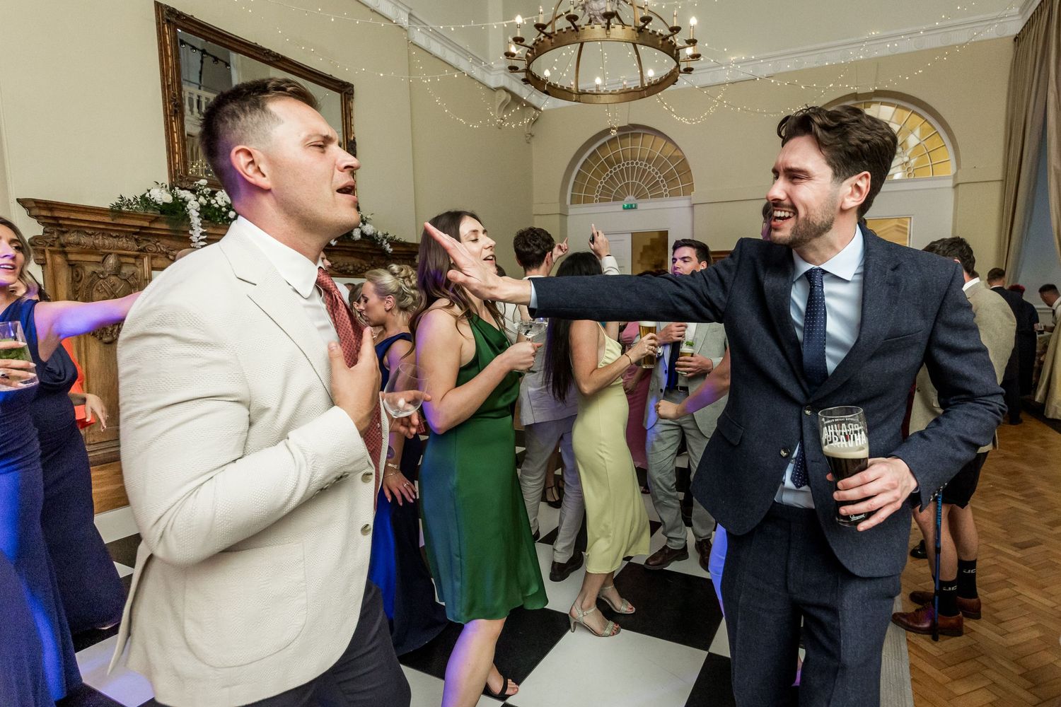 Wedding guests mingle and dance in a grand ballroom with decorative crown molding and chandeliers.
