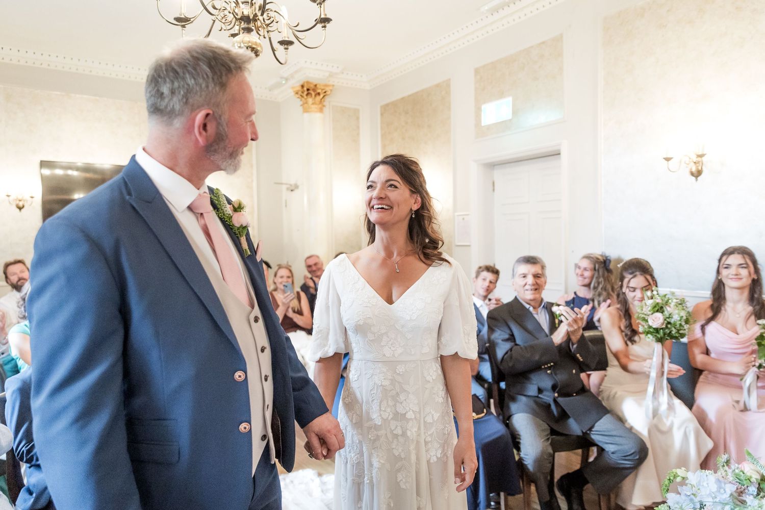 A joyful wedding ceremony moment in a bright room with guests visible in the background.