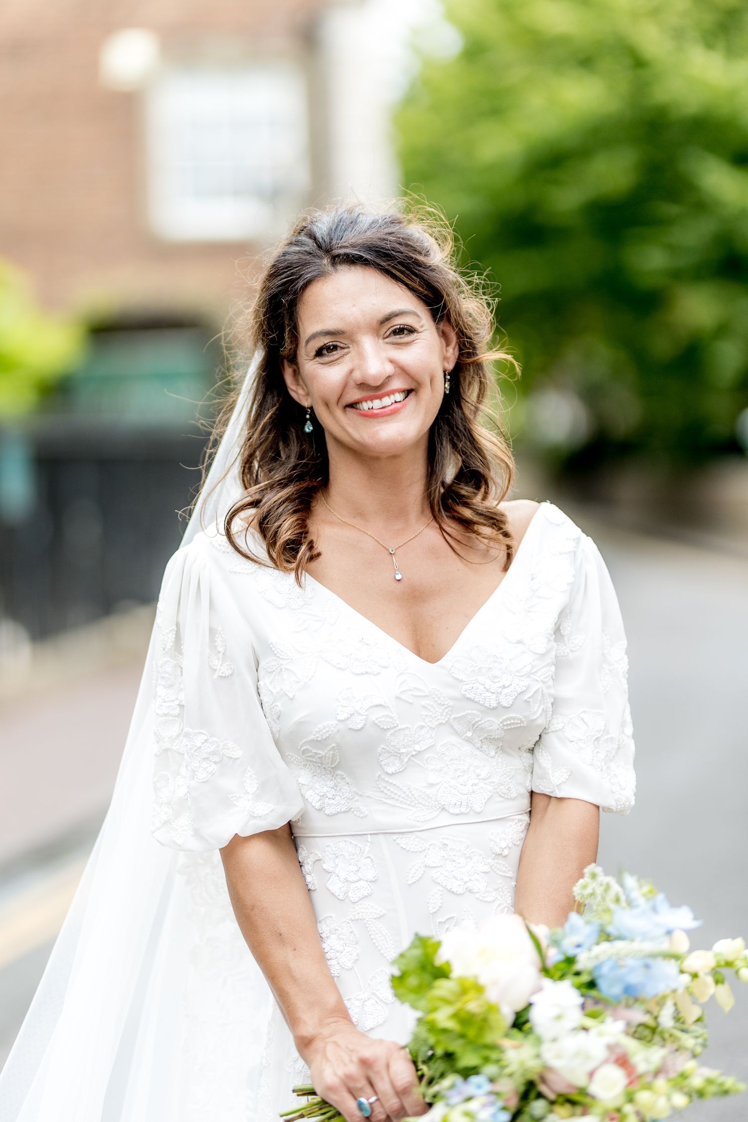 A bride in a white lace dress holds a colorful bouquet against a green garden background.