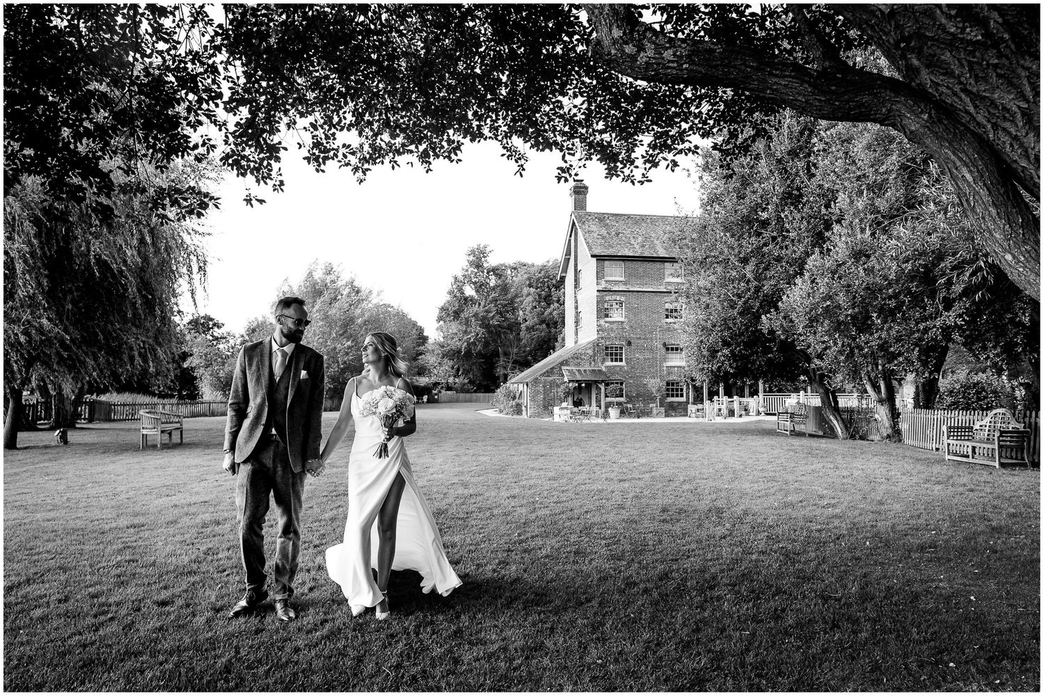 A bride and groom walk across the grounds of Sopley Mill with a view of the wedding venue in the background.