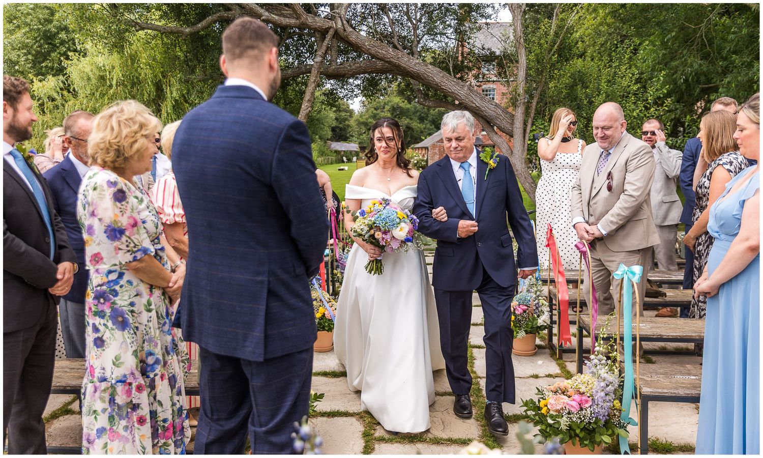 Bride walks down the aisle with her father at an outdoor wedding ceremony surrounded by guests.