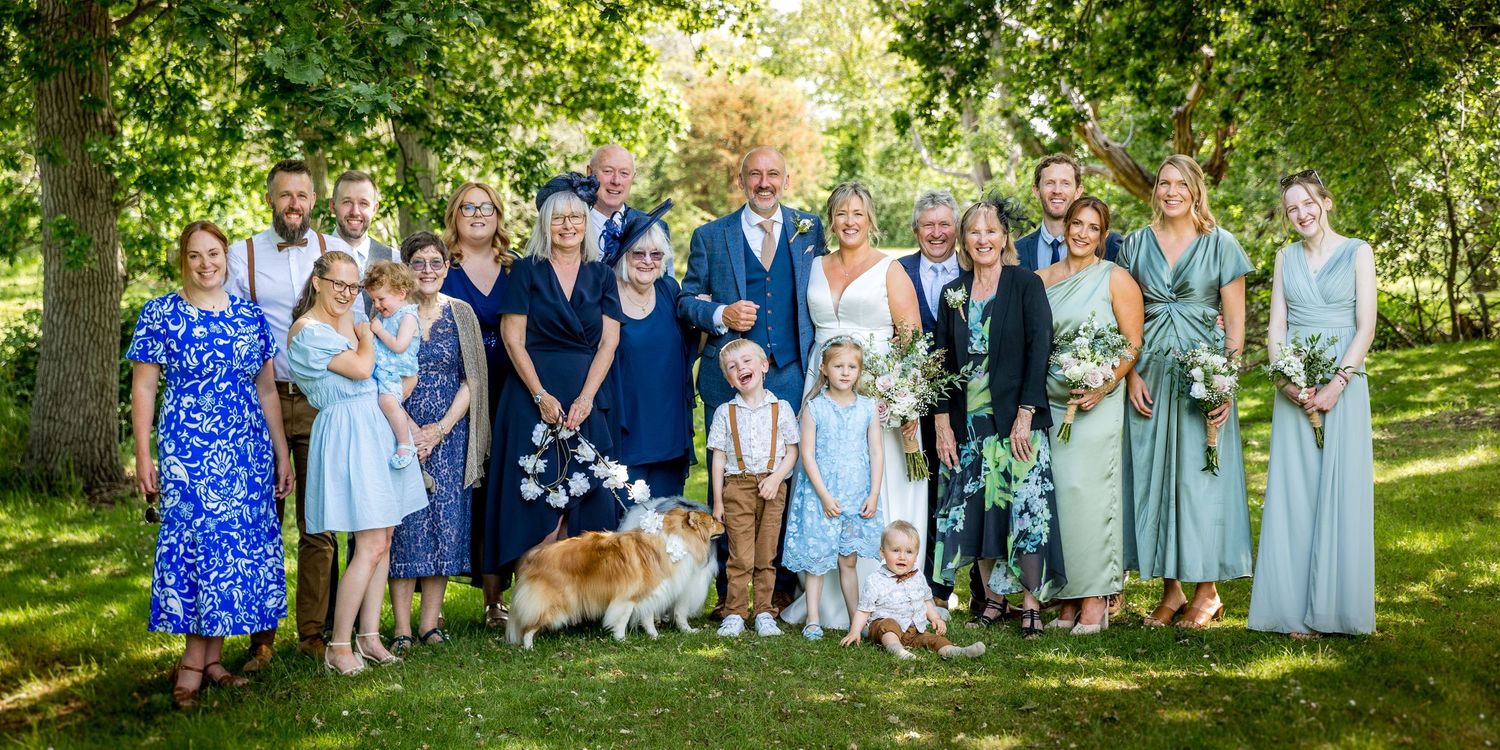 Large group wedding photo with guests gathered under green trees.