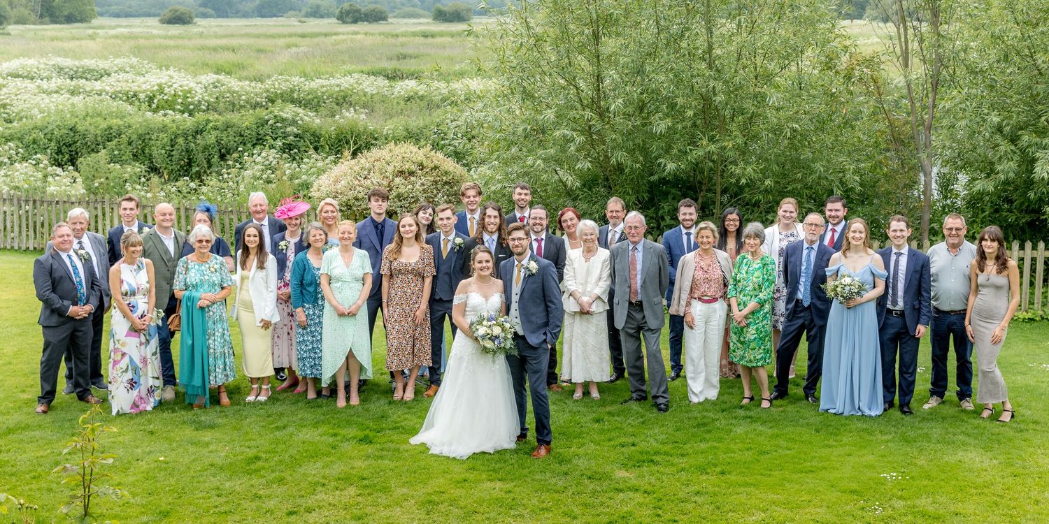 Large group wedding photo with guests lined up on grassy lawn under trees.
