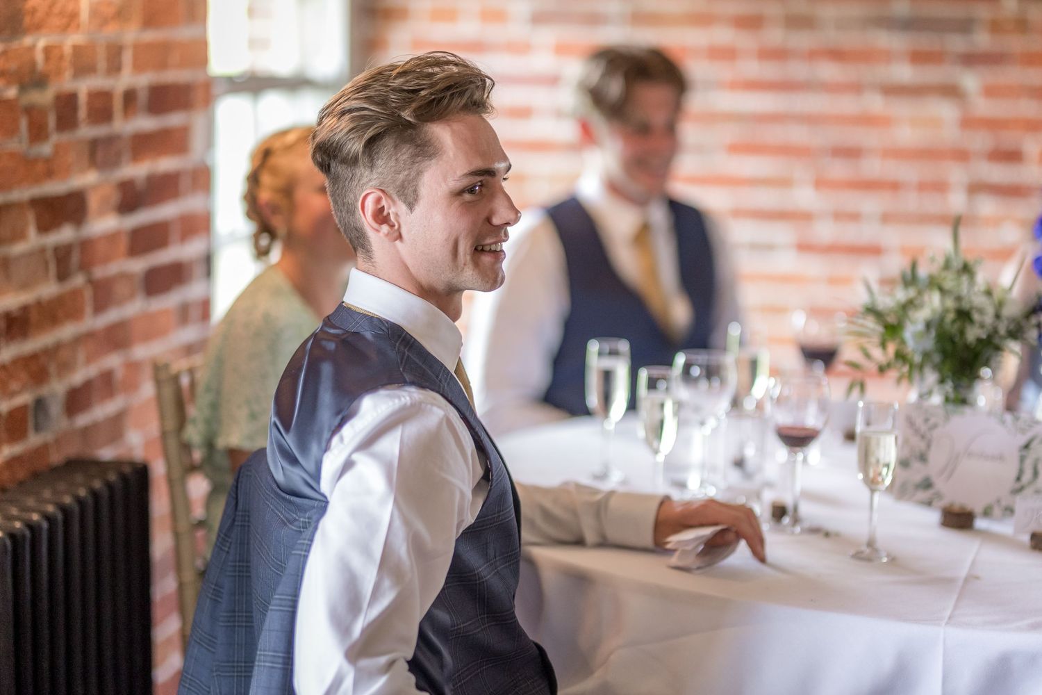 Wedding reception guests seated at tables with exposed brick walls and natural lighting in background.