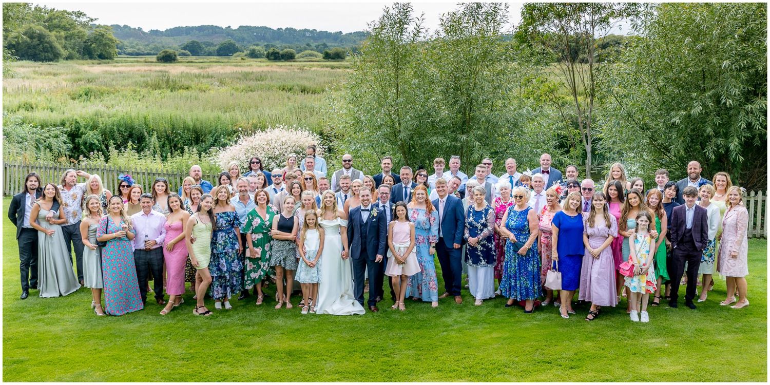 Large group wedding photo with guests arranged on grassy hillside under blue summer sky.