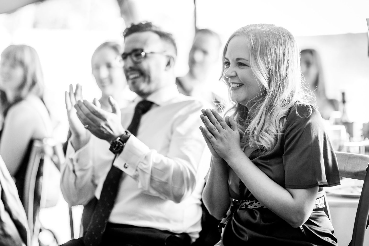 Wedding guests laugh and applaud during reception speeches in black and white.