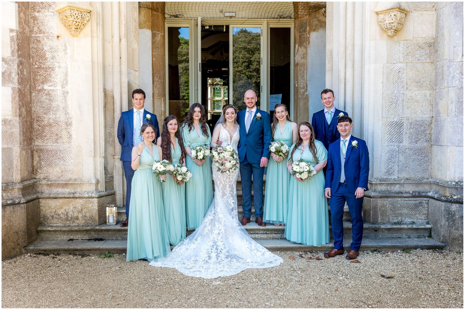 Wedding party in blue and white attire posing on the steps of a historic stone building.