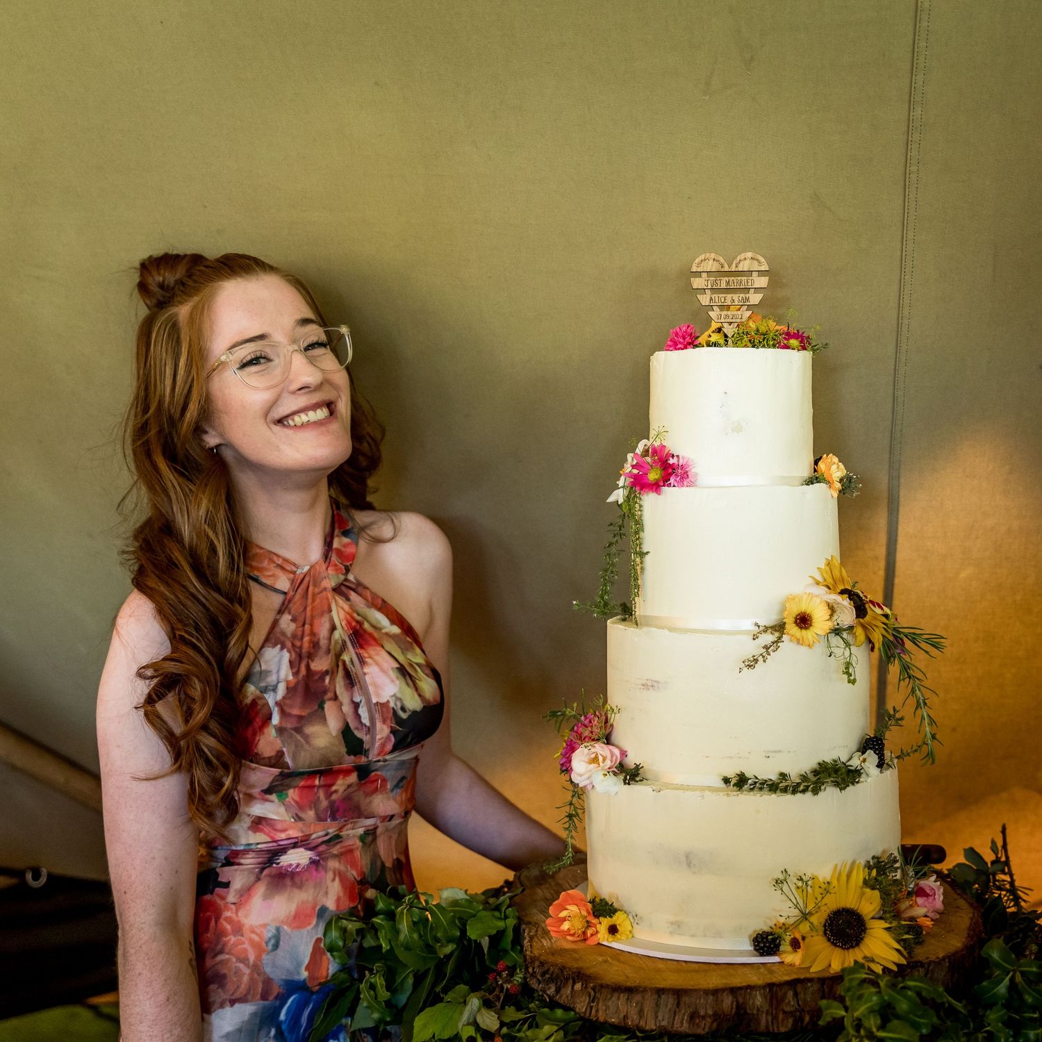 A white wedding cake decorated with sunflowers and fresh flowers on a rustic wooden stand.