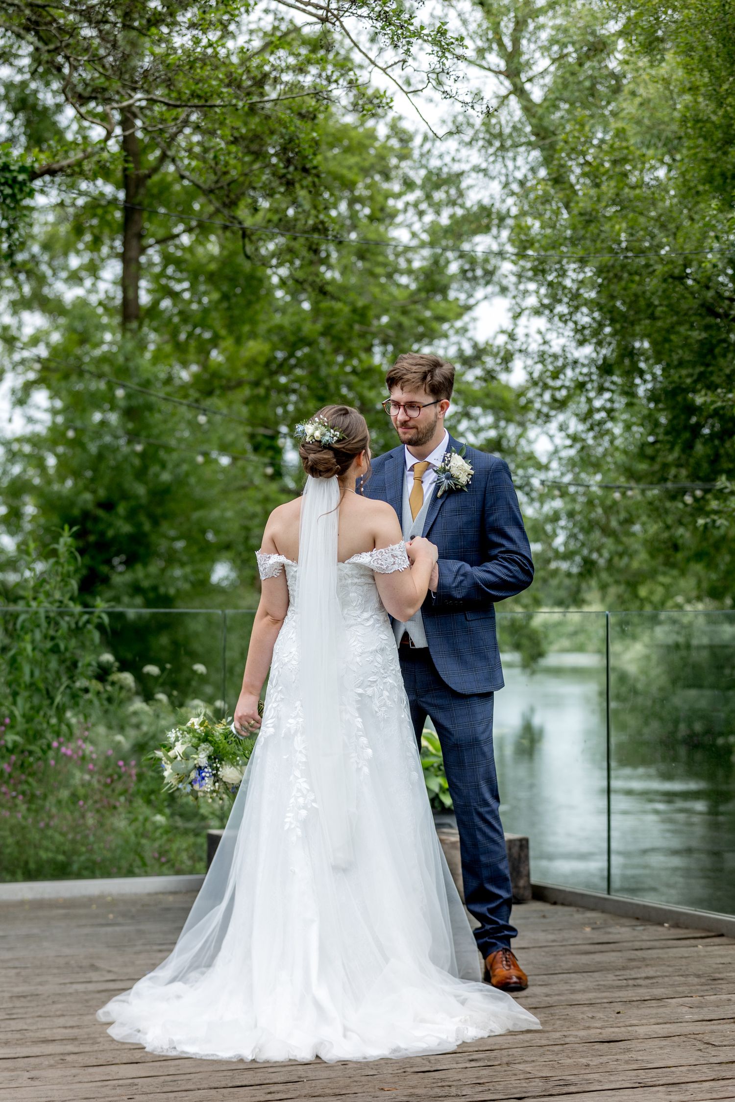 Wedding couple stands together on rustic wooden deck with glass railing and trees.