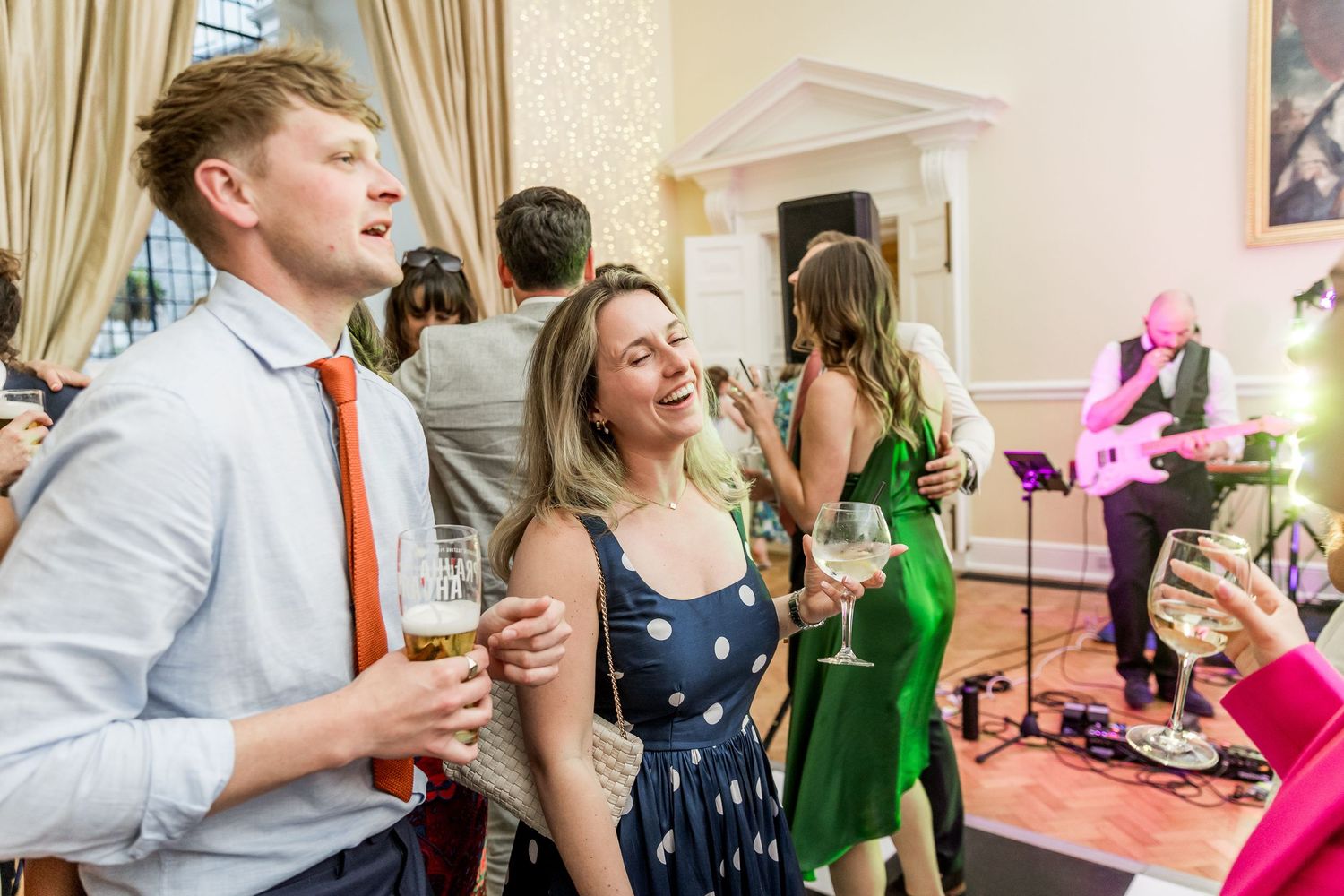 Wedding party members dance and socialize in an ornate ballroom with cream colored walls and curtains.