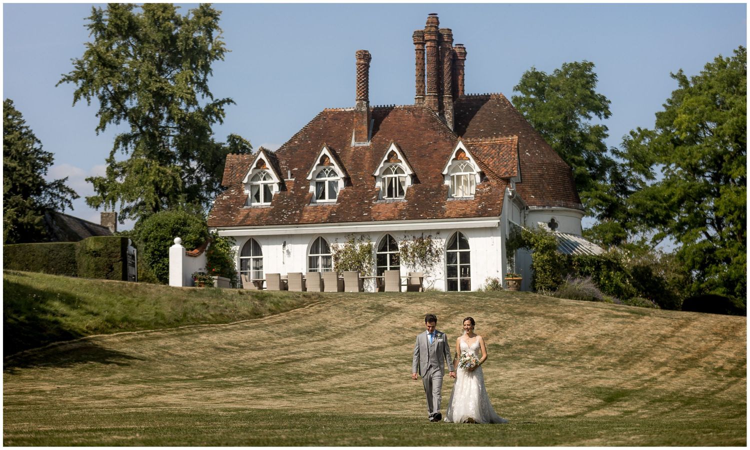 A couple stands on a hill in front of an English Tudor-style country house with distinctive chimneys.