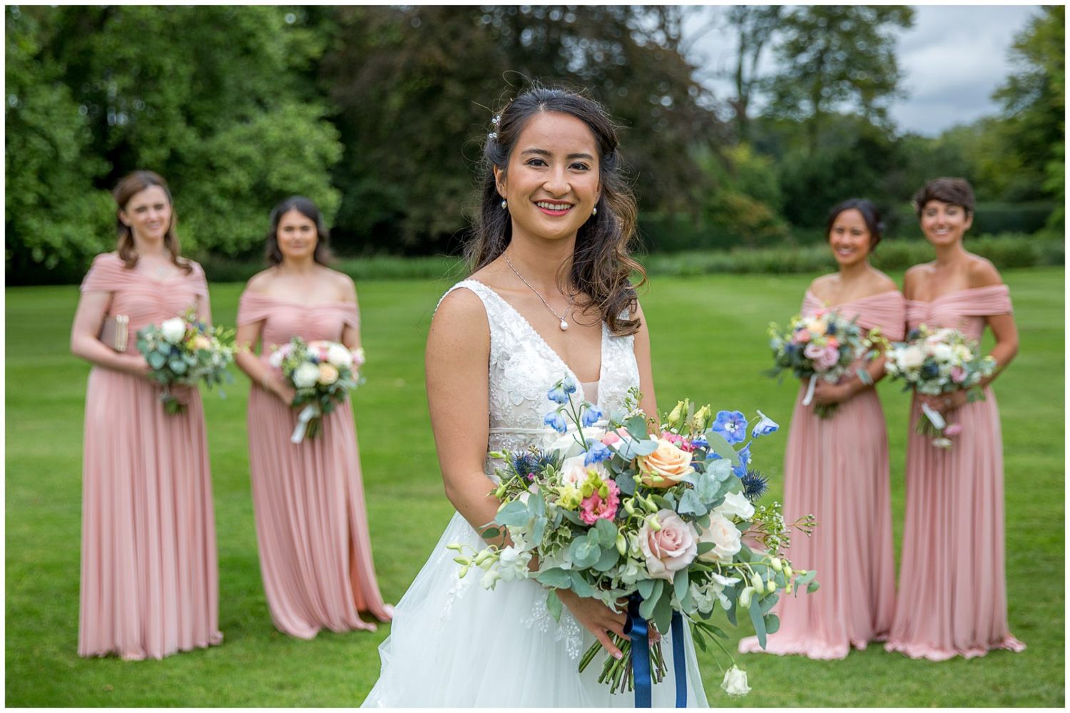 Wedding party in pink bridesmaid dresses stands on manicured lawn with floral bouquets.