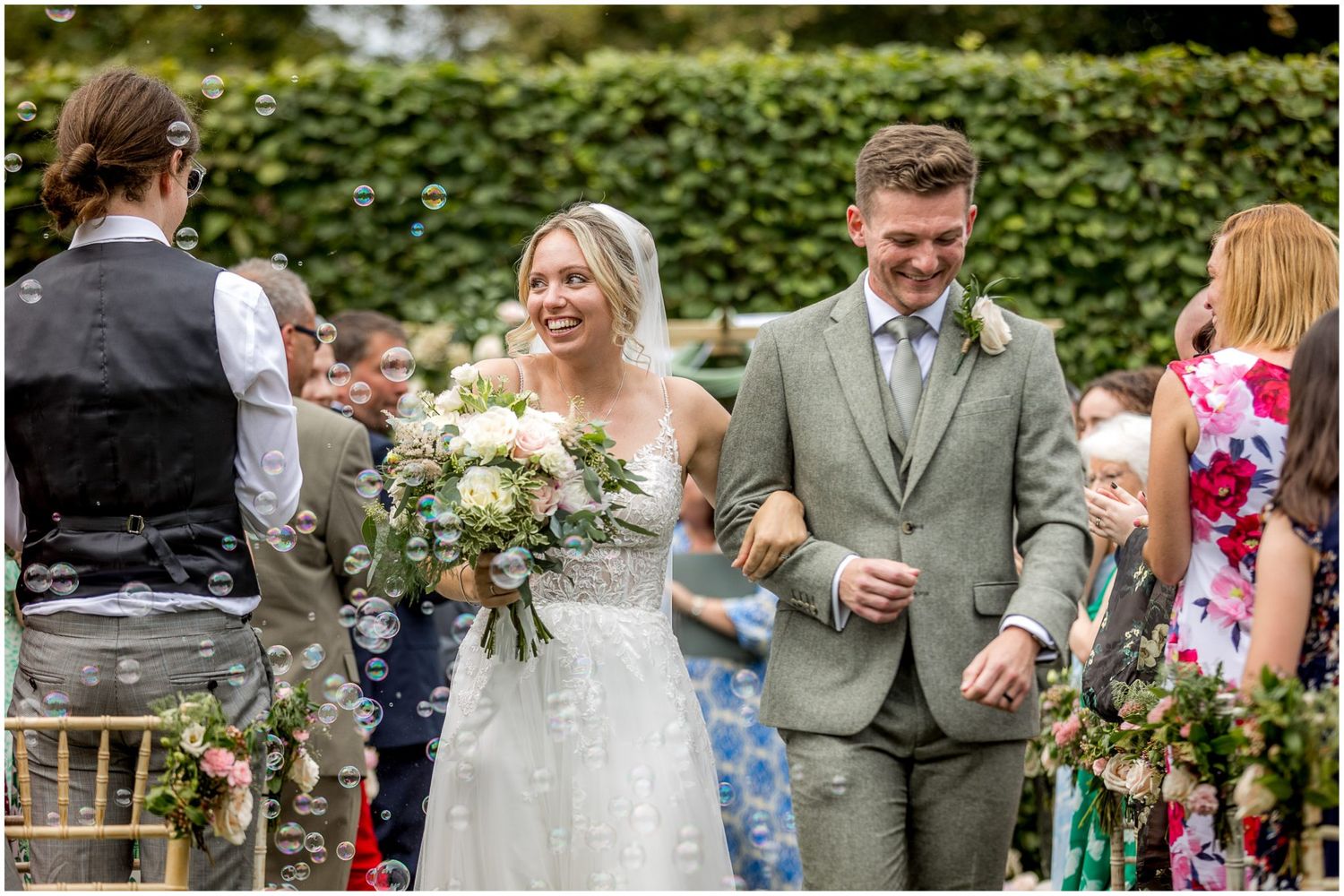 Wedding party celebrating outdoors with green foliage backdrop and decorative floral arrangements.