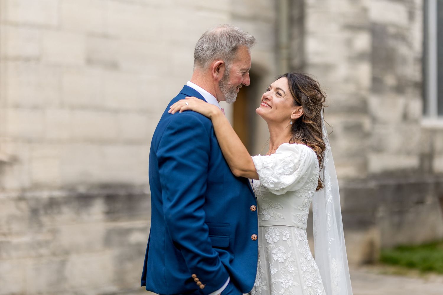 Elegant wedding couple poses together against stone wall backdrop in romantic outdoor setting.