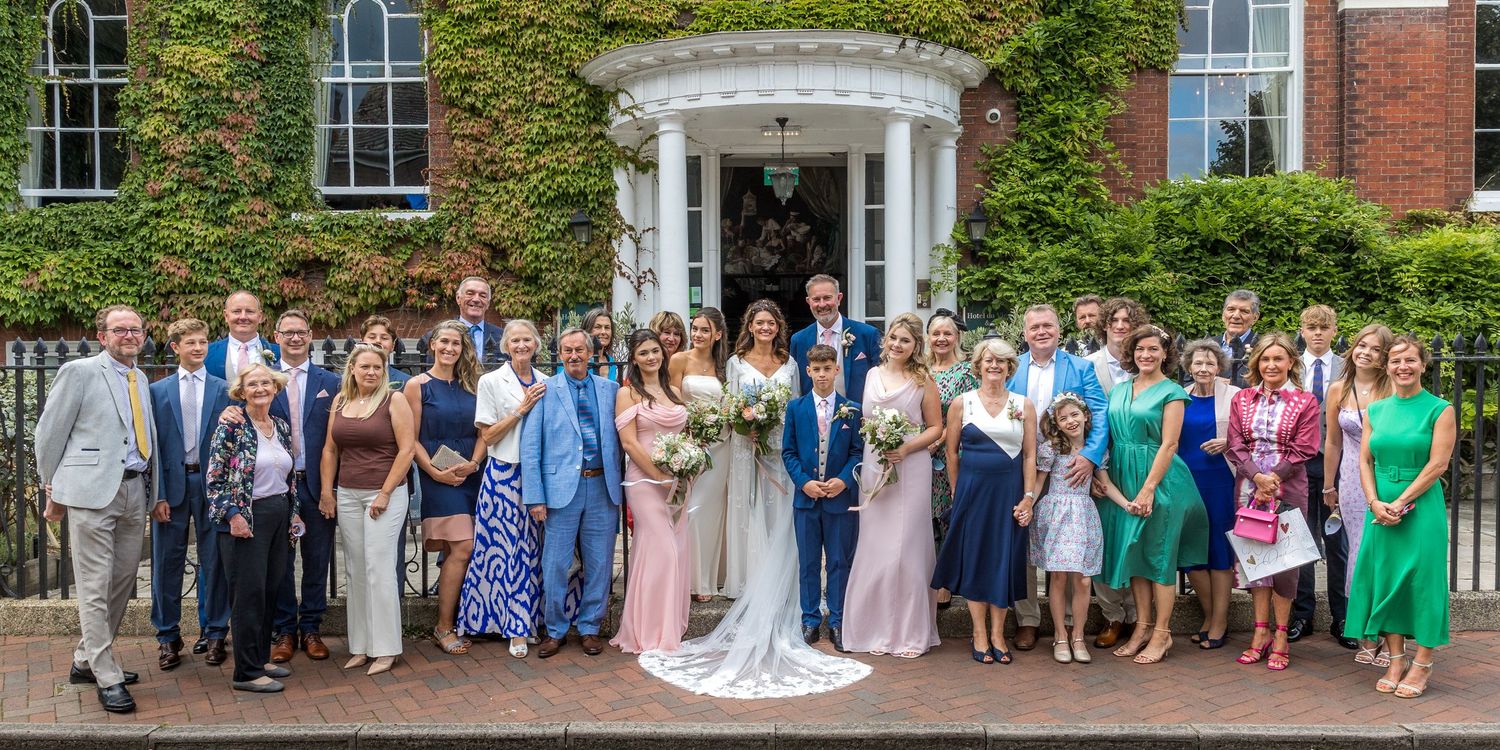 Large wedding party group photo taken outside historic brick building with ivy-covered walls.