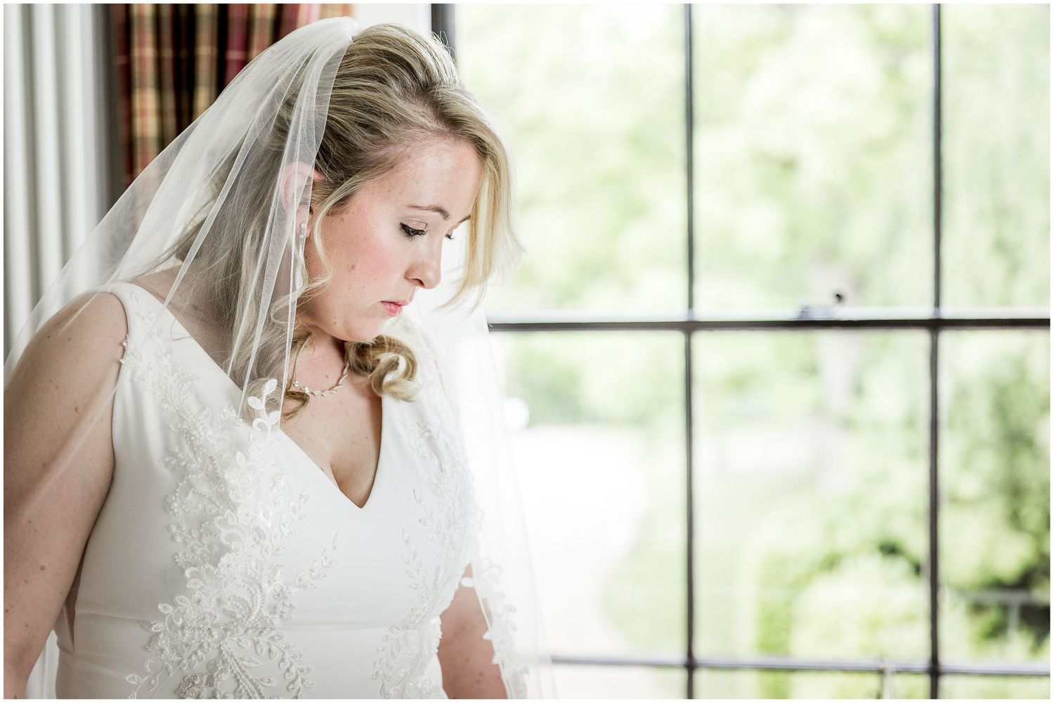 Elegant bride in a lace wedding dress poses by a bright window in a classic indoor setting.