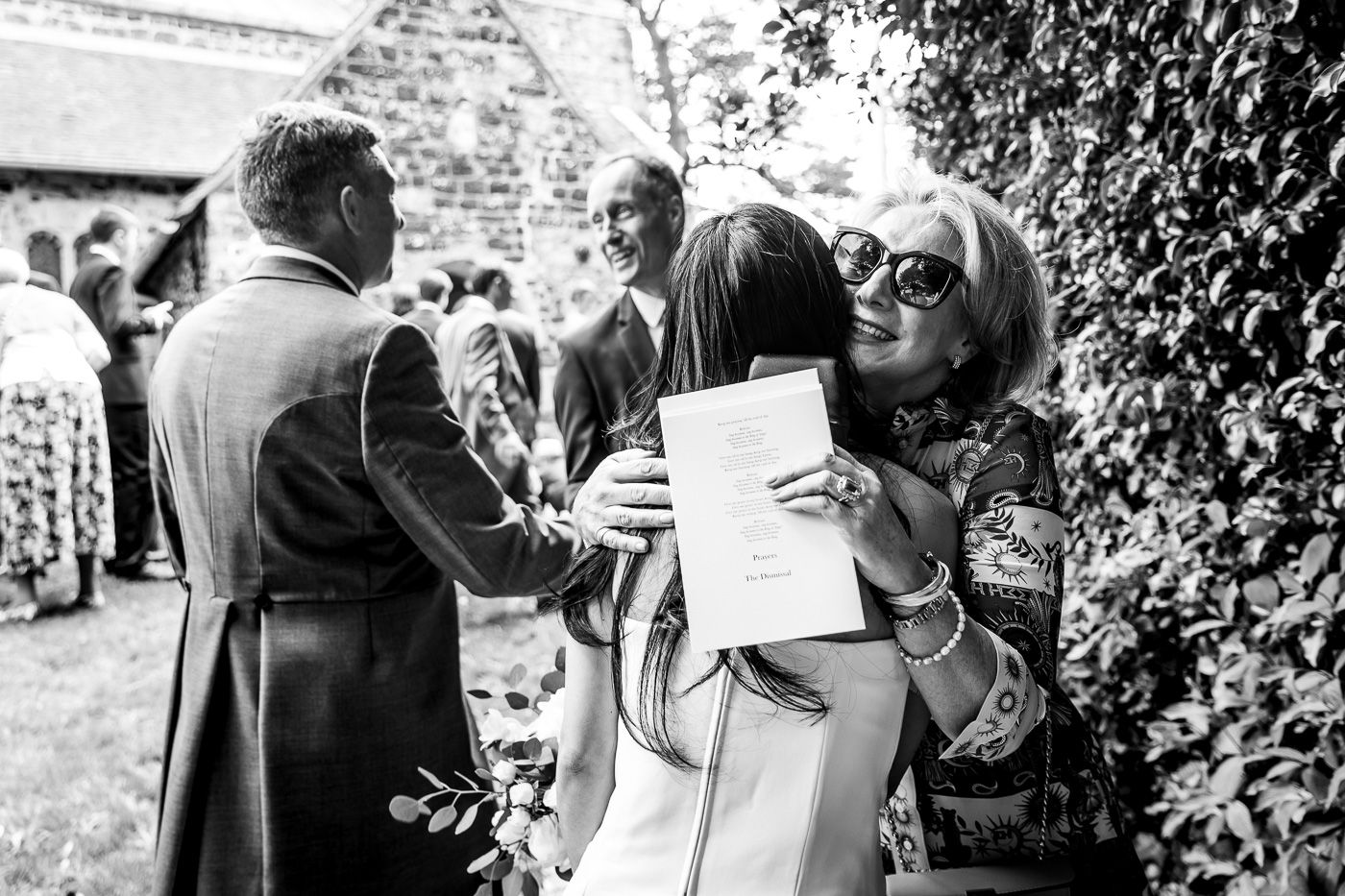 Black and white candid photo of wedding guests greeting and hugging at an outdoor reception.