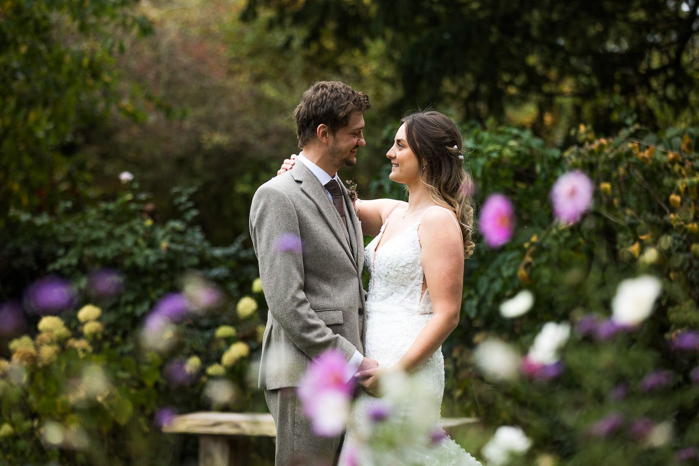 Newlyweds embrace in beautiful garden setting with purple flowers in foreground.