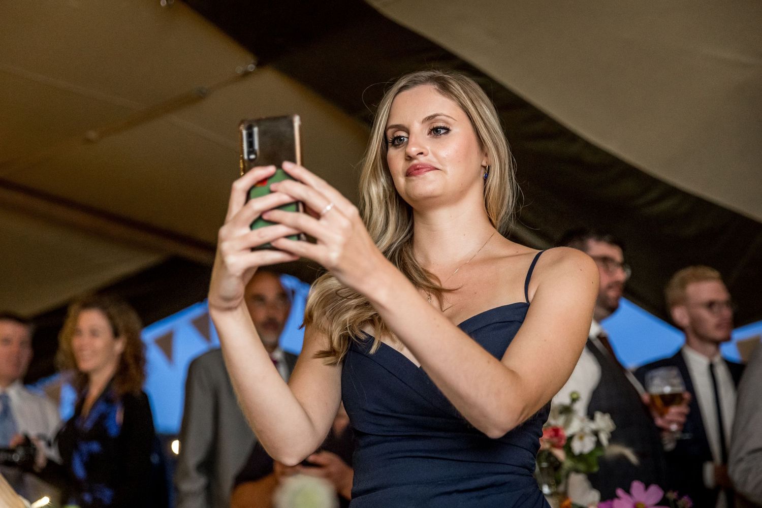 A guest in a navy dress takes a photo with their phone during a wedding reception.