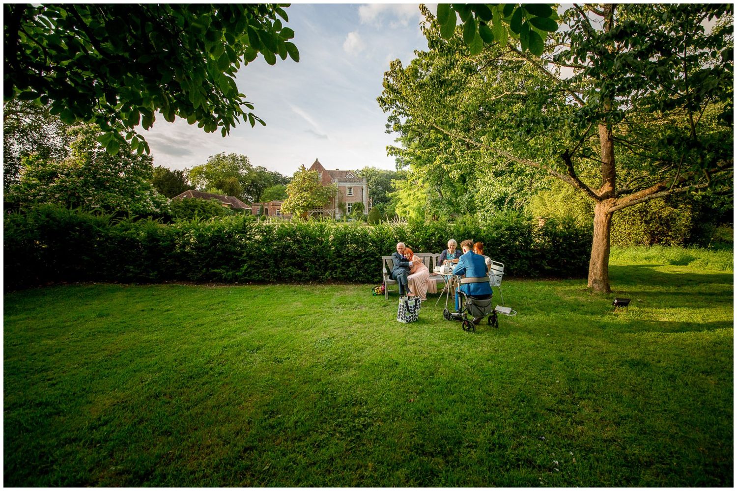 People enjoying a sunny day in a lush green park with scattered trees and rolling hills in the background.