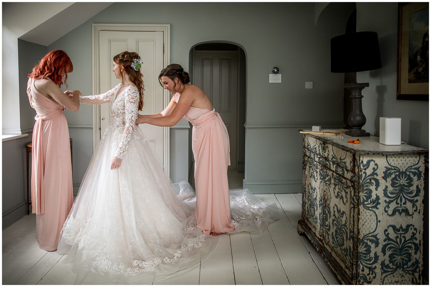 Two bridesmaids in blush pink dresses help adjust a bride's elegant white lace wedding gown in a grey room.