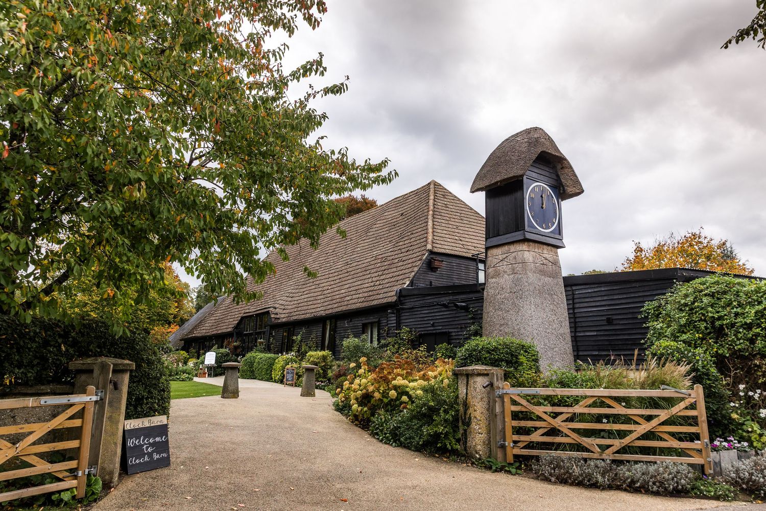 Historic black wooden barn with thatched roof stands among autumn trees on a cloudy day in the English countryside.