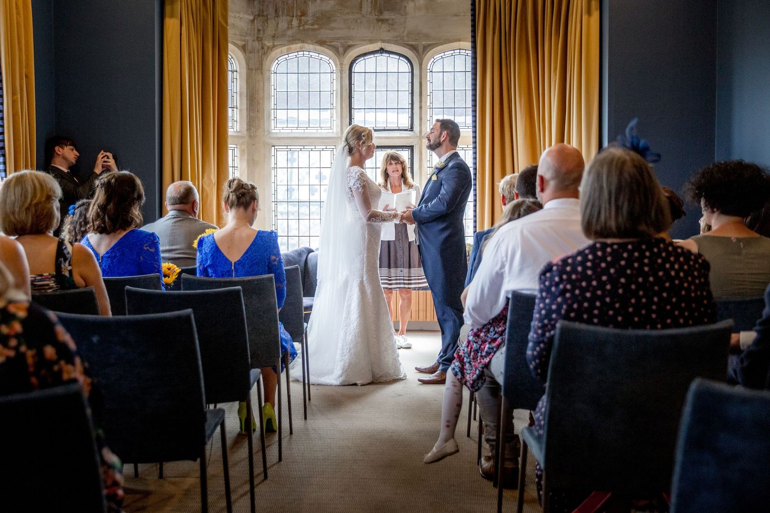 A wedding ceremony in progress in a room with large arched windows and golden drapes.