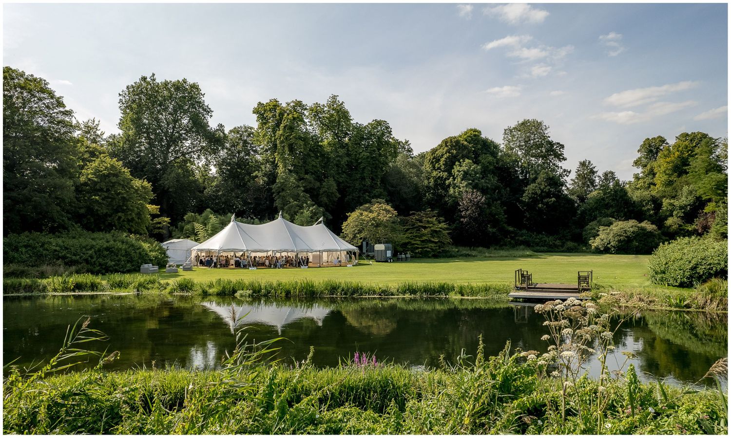 A serene view of a white wedding tent reflected in a pond surrounded by lush greenery.