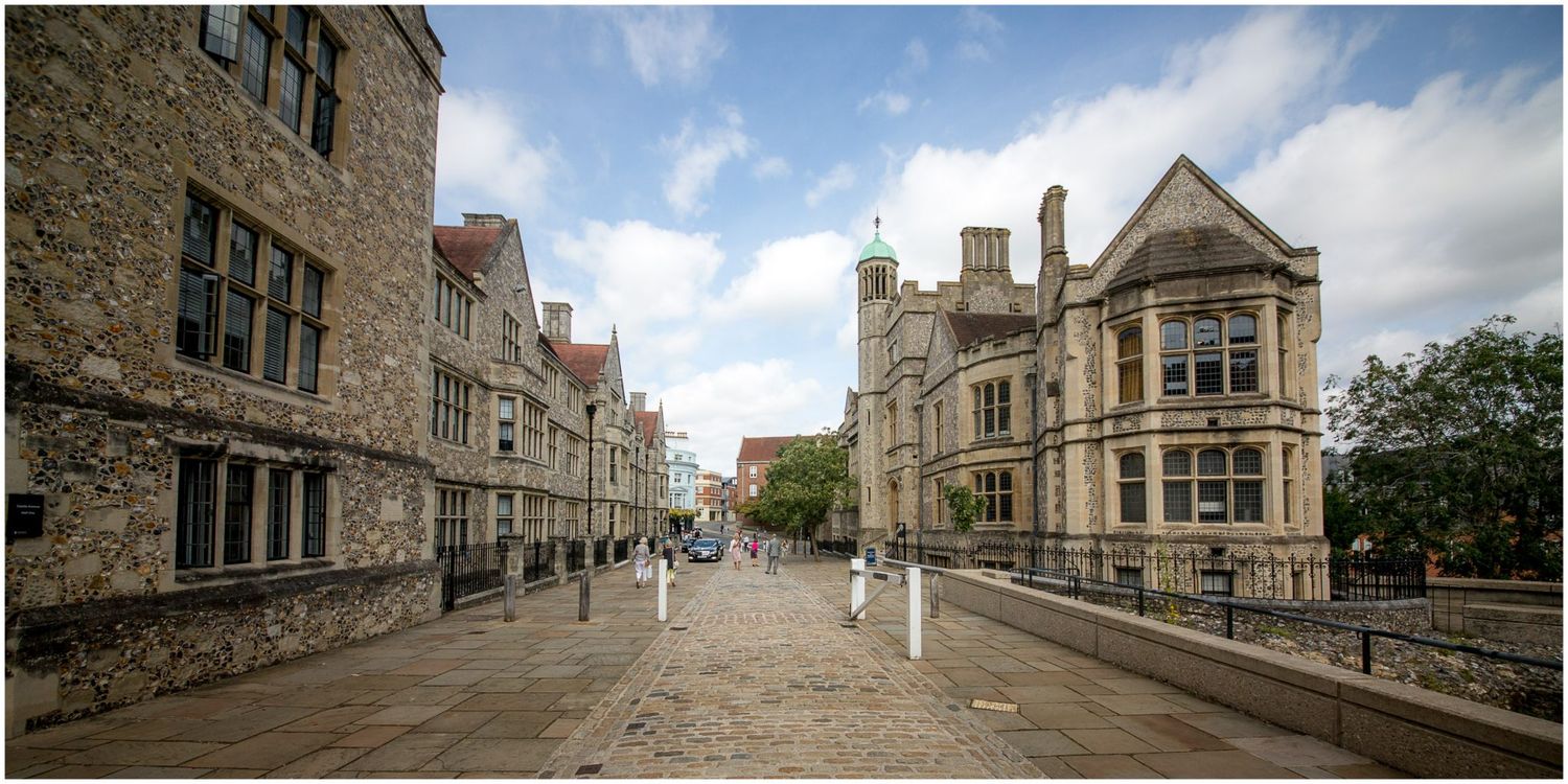 Historic stone buildings with Gothic architecture line a cobblestone street under cloudy skies at Oxford University.