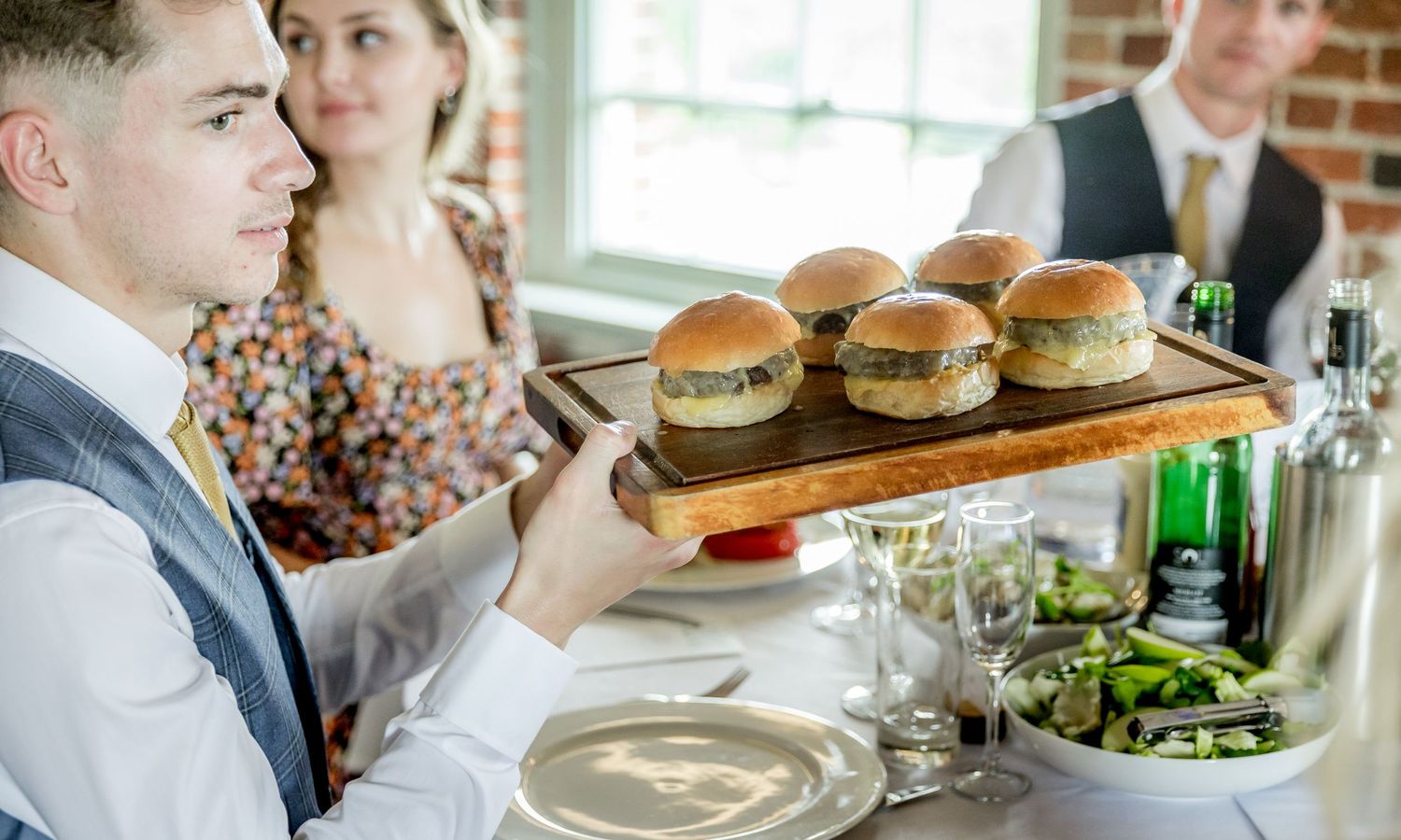 Wedding reception featuring casual dining with sliders and salads served on rustic wooden boards.