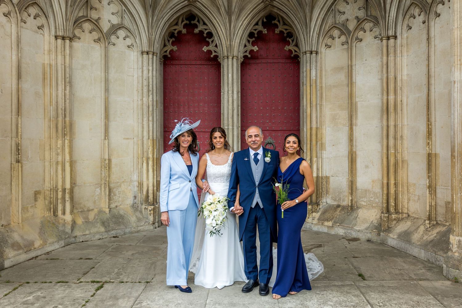 Wedding party poses in formal attire against ornate Gothic stone architecture and columns.