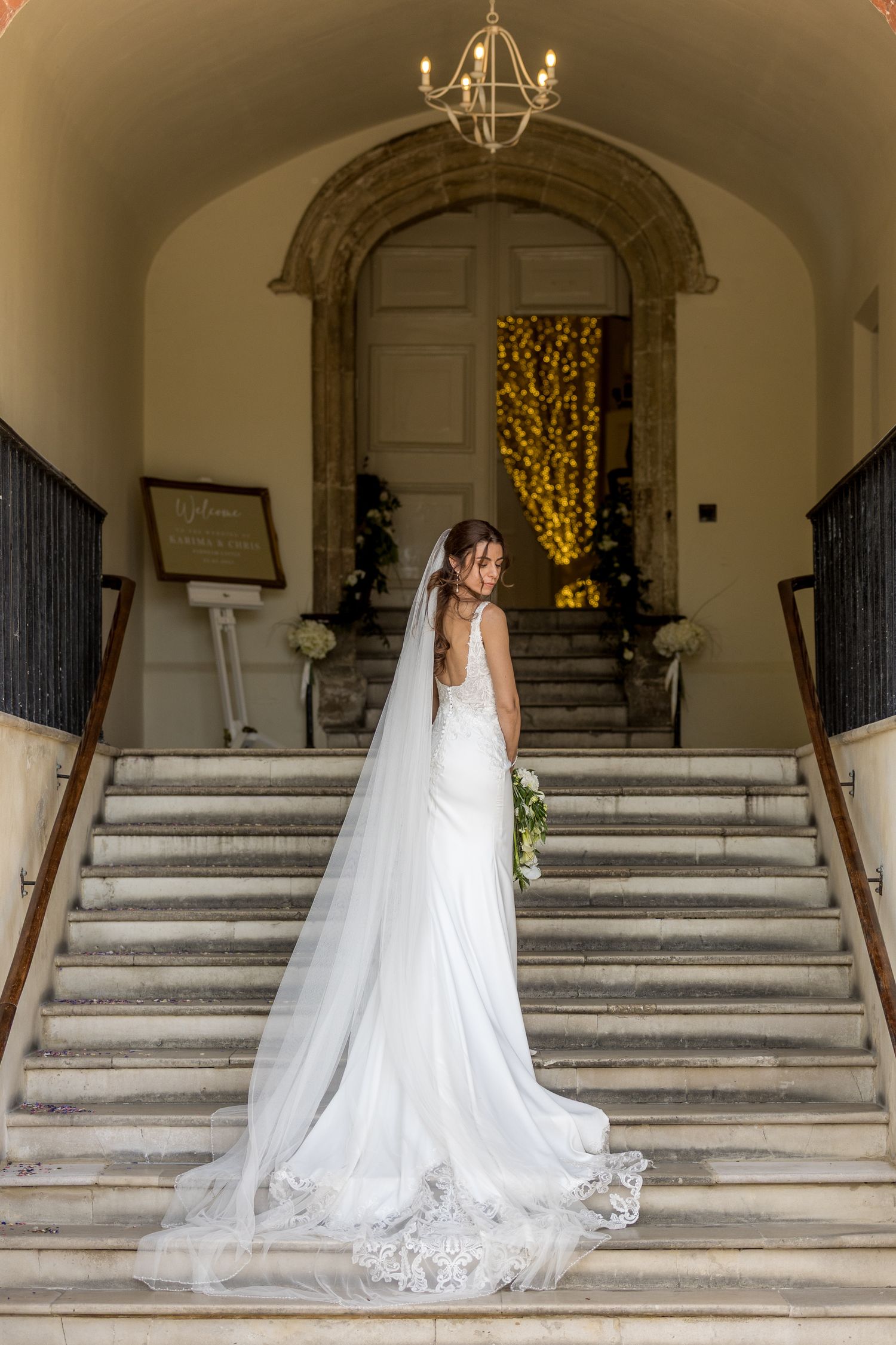Bride in flowing lace gown stands gracefully on marble staircase with long train trailing behind.