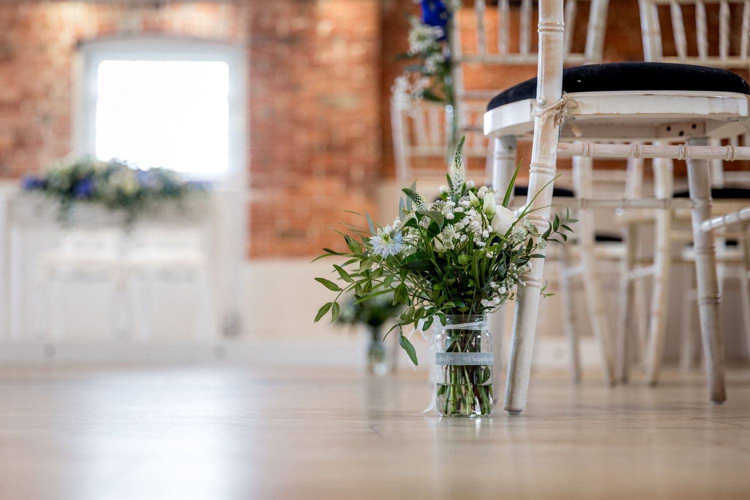 Wedding reception setup with white chairs and floral centerpieces against brick walls.