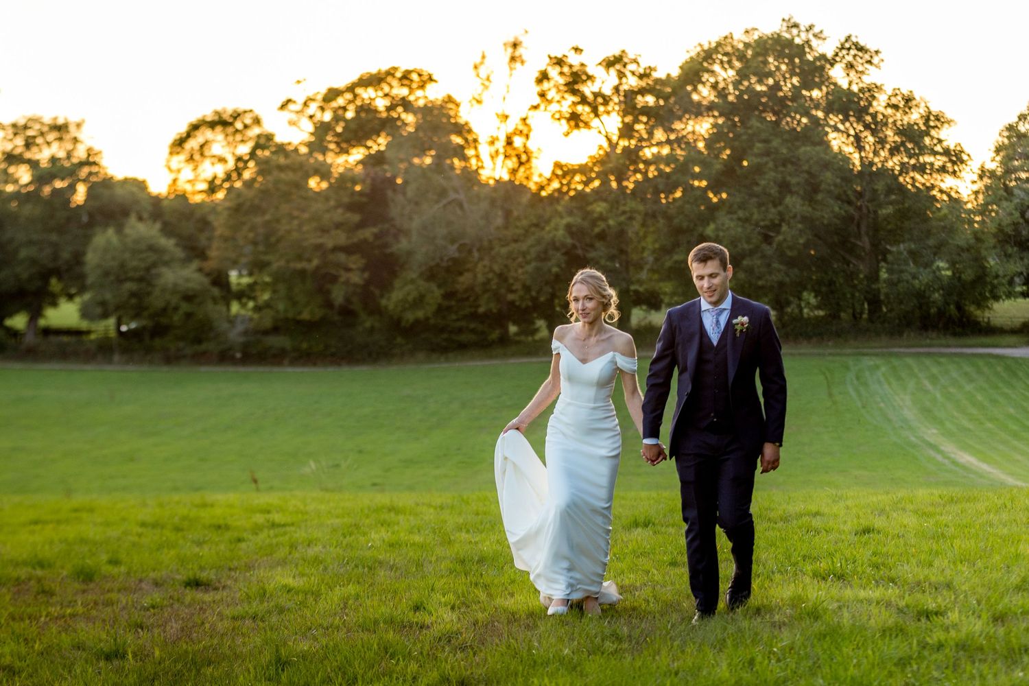 A couple walks through a golden meadow during sunset with dramatic sky overhead.