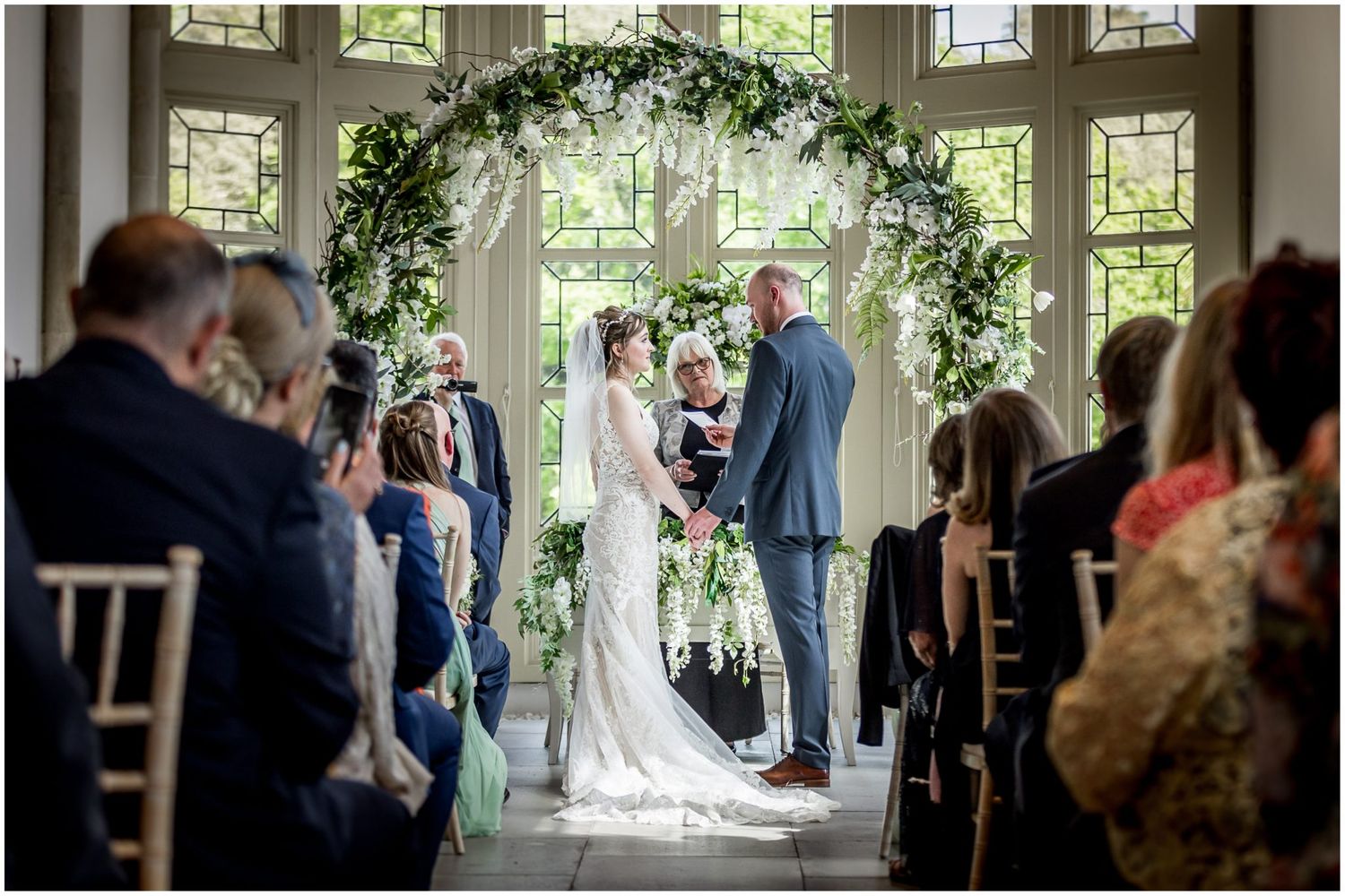 Indoor wedding ceremony setup with greenery arch and natural window lighting.