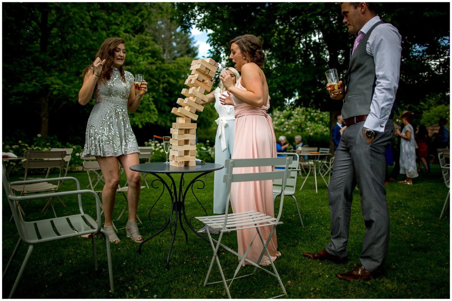 People gather outdoors for a garden party with a giant Jenga game and bistro tables on a summer afternoon.