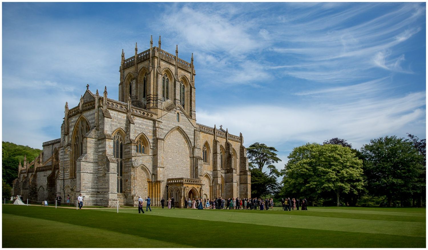 Historic Milton Abbey church and grounds in Dorset features Gothic architecture against a bright blue summer sky.